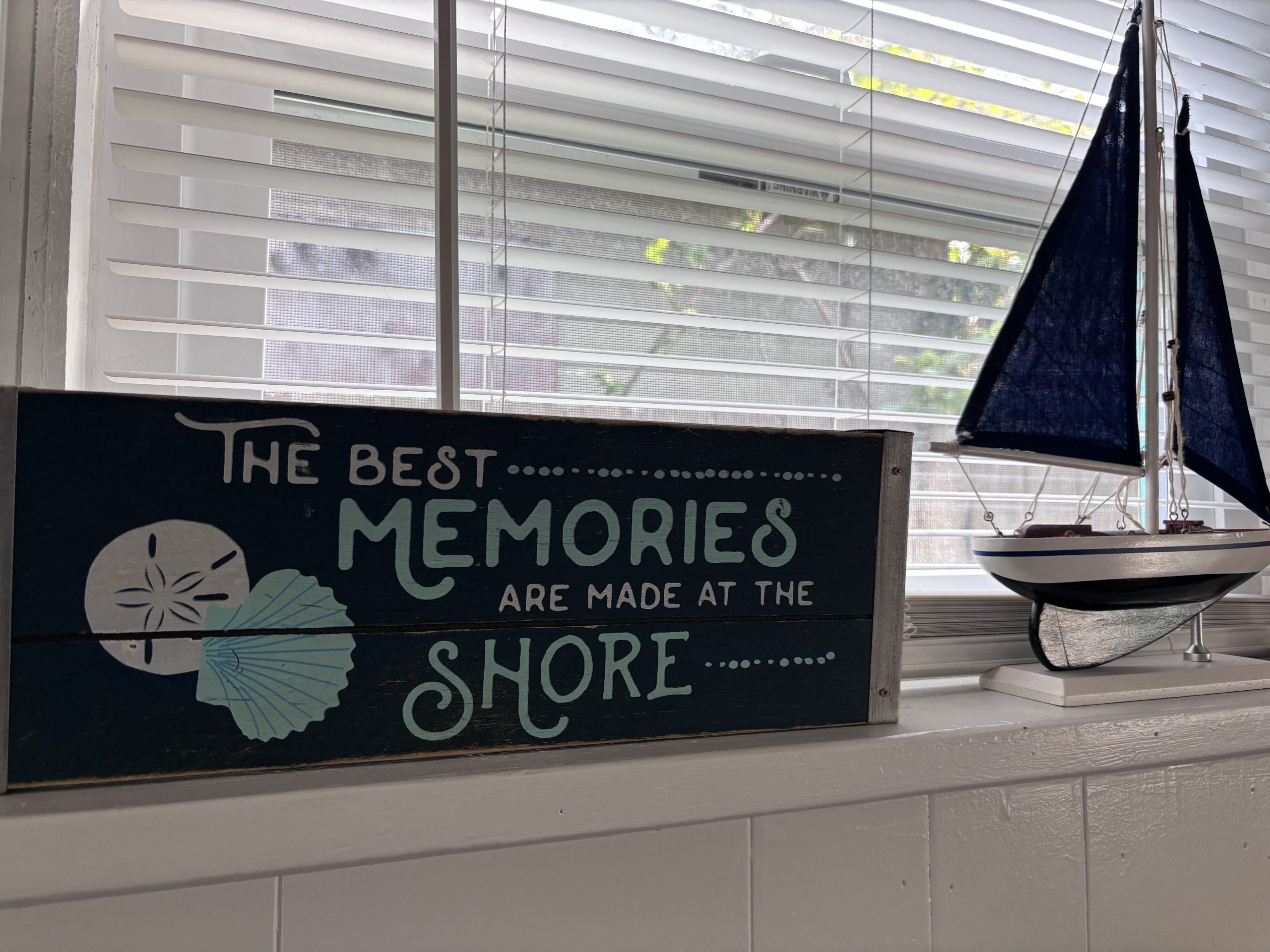 A decorative wooden sign on a windowsill reads “The best memories are made at the shore” with illustrations of a sand dollar and scallop shell. Beside it sits a small model sailboat with navy blue sails, and white blinds cover the window behind them.