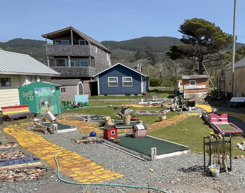 Whimsical roadside miniature golf course in a beach town, with colorful hand-painted features including a yellow brick road, benches, and Wizard of Oz mural; coastal mountains and eclectic beach homes in the background.