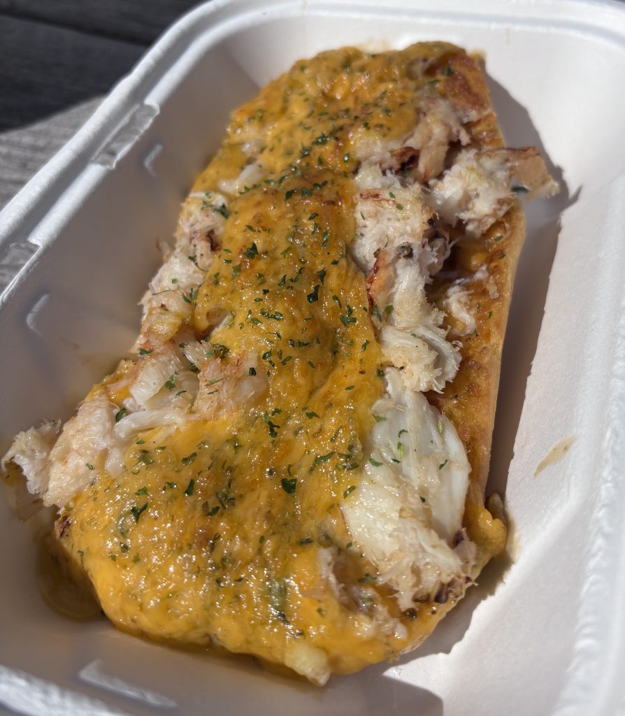 Close-up of a cheesy open-faced crab melt sandwich in a white compostable takeout container, with chunks of crab meat and melted golden cheddar, topped with herbs, photographed outdoors in natural light.