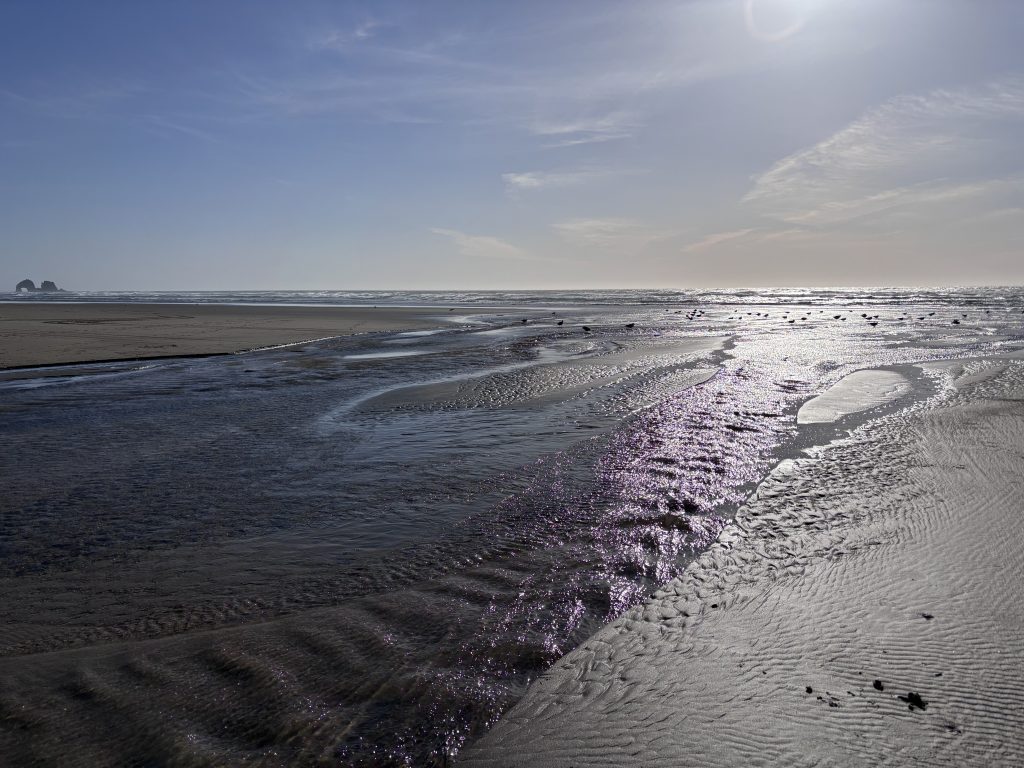 A shimmering tidal stream flows across a flat, sandy beach toward the ocean under a clear blue sky. The sun is low on the horizon, creating dramatic reflections on the water. In the distance, a rock formation with two arches rises from the sea.