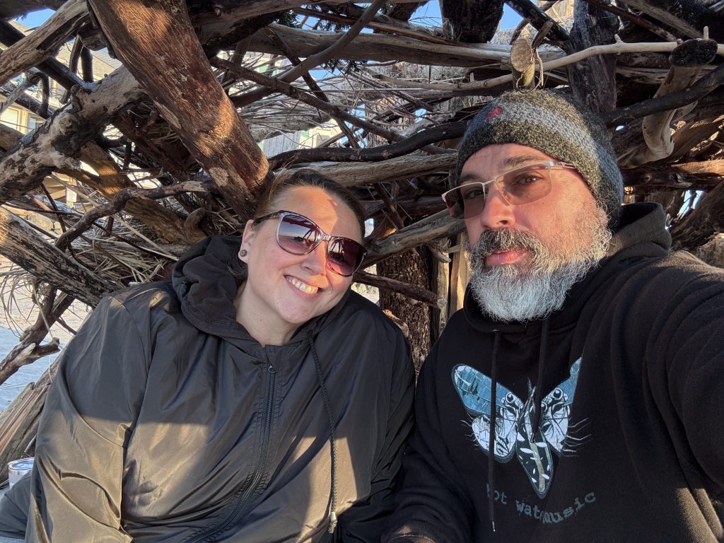 Friends sitting under a cozy driftwood fort on the beach; the woman is smiling brightly in sunglasses and a warm jacket, while the man wears a knit beanie and hoodie with a white beard and serious expression.