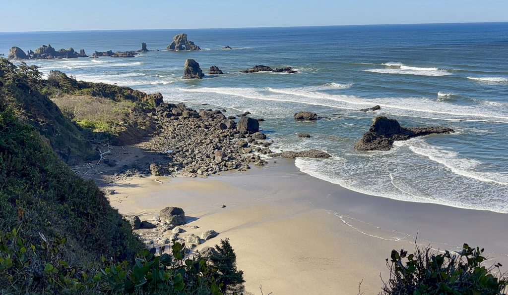 Scenic coastal view from Ecola State Park with rugged sea stacks, rocky shoreline, and waves crashing against the Oregon coast under a bright blue sky.