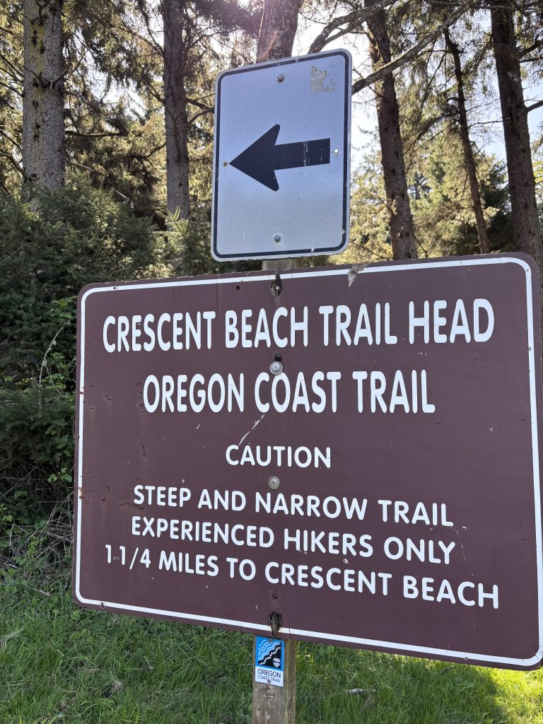 Trailhead sign for Crescent Beach on the Oregon Coast Trail warning of a steep and narrow trail suitable for experienced hikers, surrounded by dense forest.