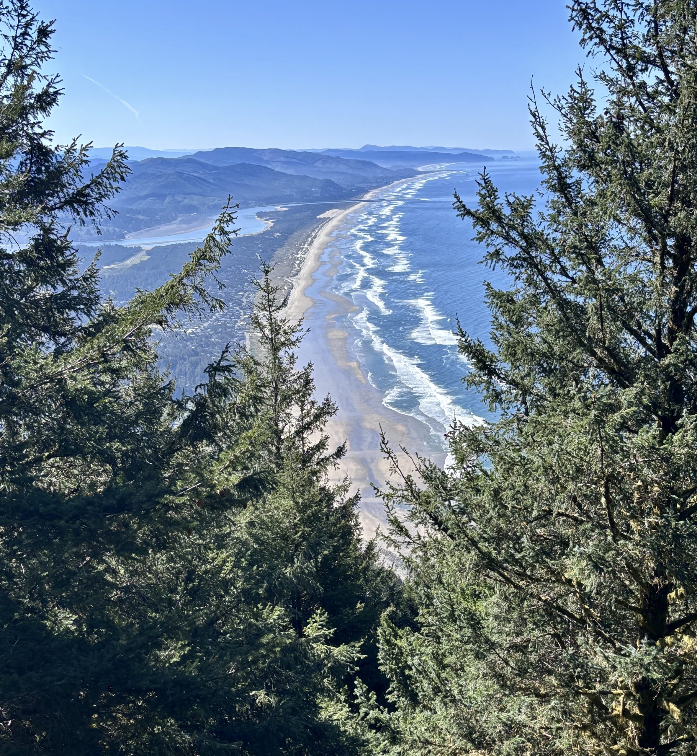 A dramatic vista from a high vantage point at the summit of Neahkahnie Mountain reveals a long, sandy beach stretching along the coastline. The turquoise ocean merges with the horizon, while a chain of distant mountains forms a scenic backdrop. Lush greenery frames the view from above.