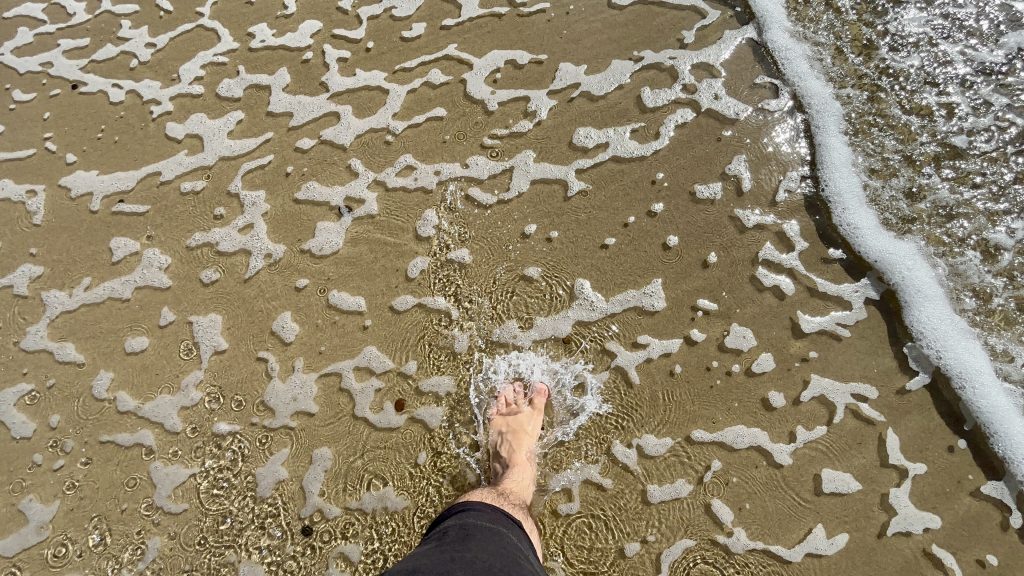 A close-up view of a sandy beach with a person's foot immersed in shallow water. The receding wave creates a foamy pattern on the sand, capturing a moment of interaction with the ocean.