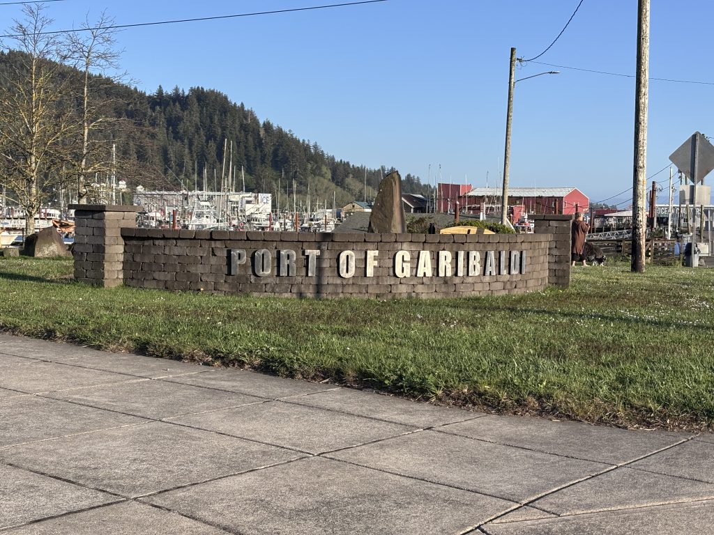 A brick and stone monument reads 'Port of Garibaldi' against a backdrop of a waterfront town. Buildings line the harbor, and lush green hills rise in the distance.