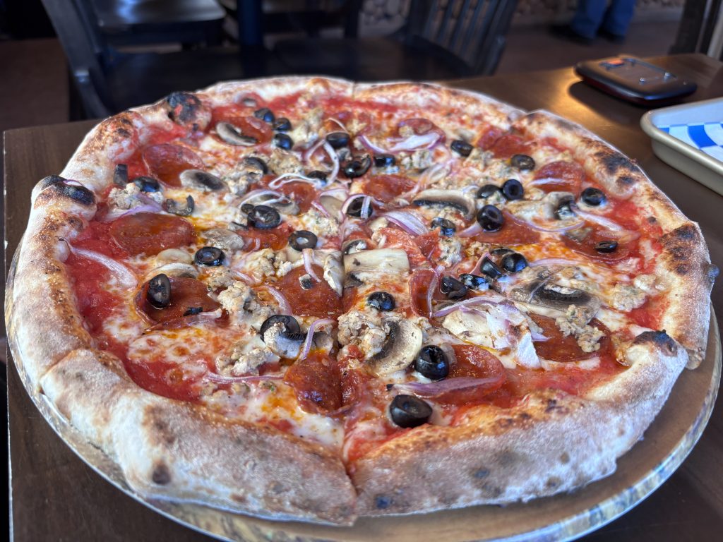 A close-up of a pizza on a metal plate, showcasing a variety of toppings including black olives, red peppers, mushrooms, and red onion, set against a red tomato sauce base.