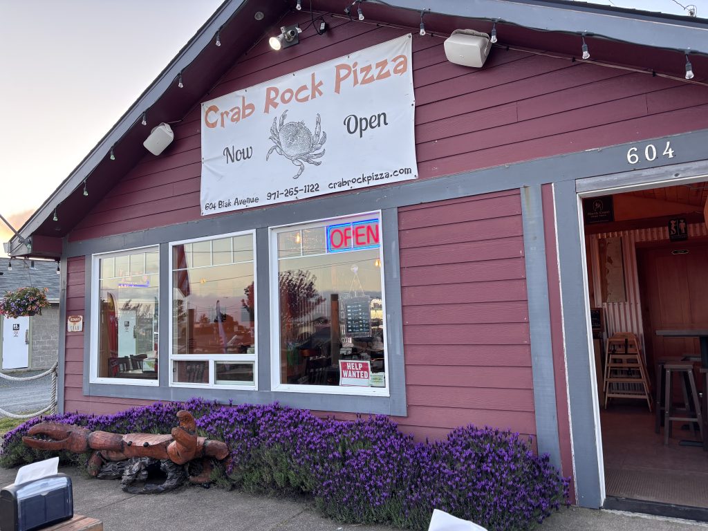 The exterior of a restaurant, 'Crab Rock Pizza,' displayed with a sign boasting its open status and contact information. Purple flowers line the front of the building.