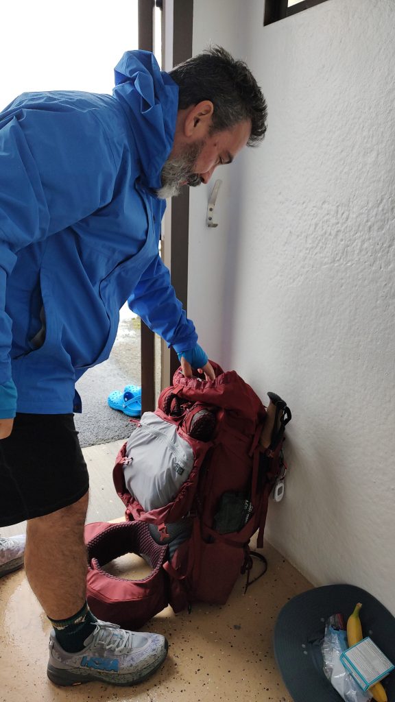 Backpacker in blue rain jacket preparing gear indoors with red Osprey pack, HOKA trail shoes, and hiking essentials — Oregon Coast Trail thru-hike prep.