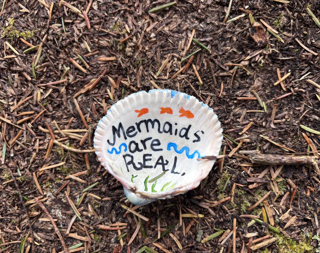 Painted seashell with the words "Mermaids are real" lying on forest floor covered in pine needles and moss along the Oregon Coast Trail.