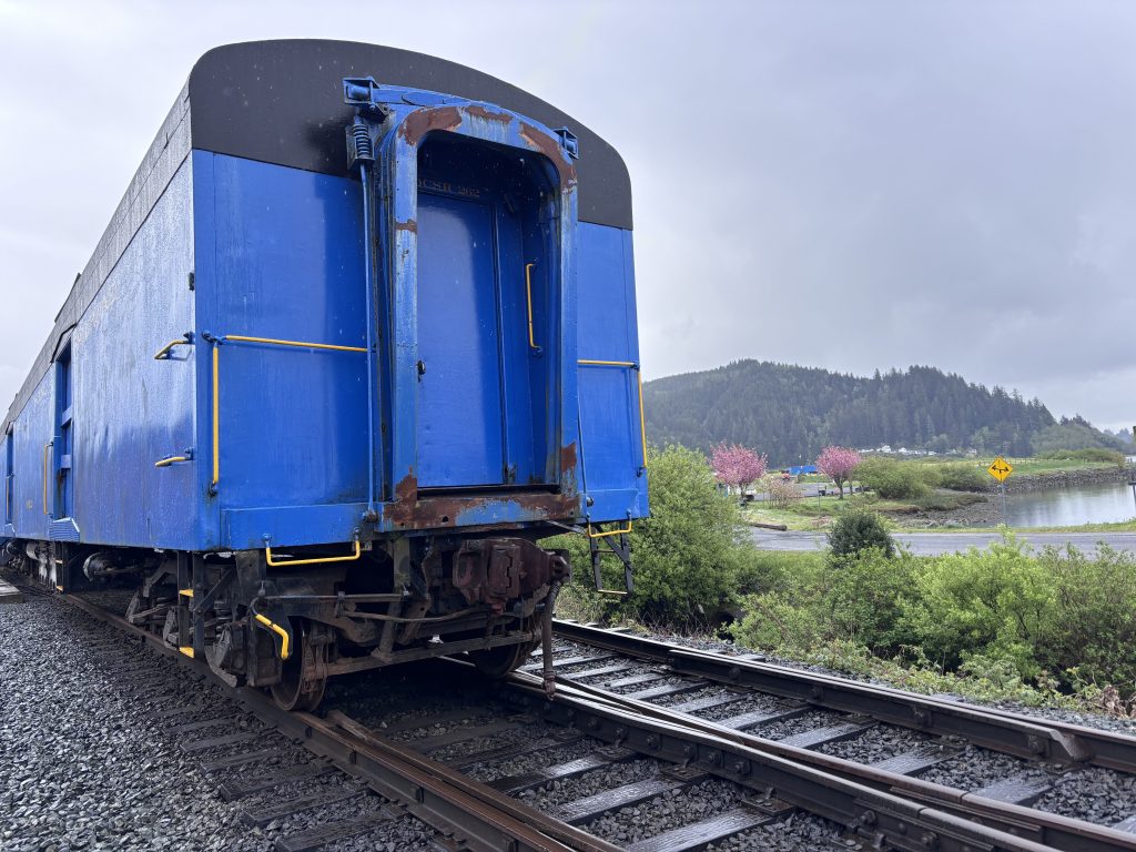 3. Bright blue train car on railroad tracks near a marshy area and forested hills under gray cloudy skies at the Oregon Coast Scenic Railroad.