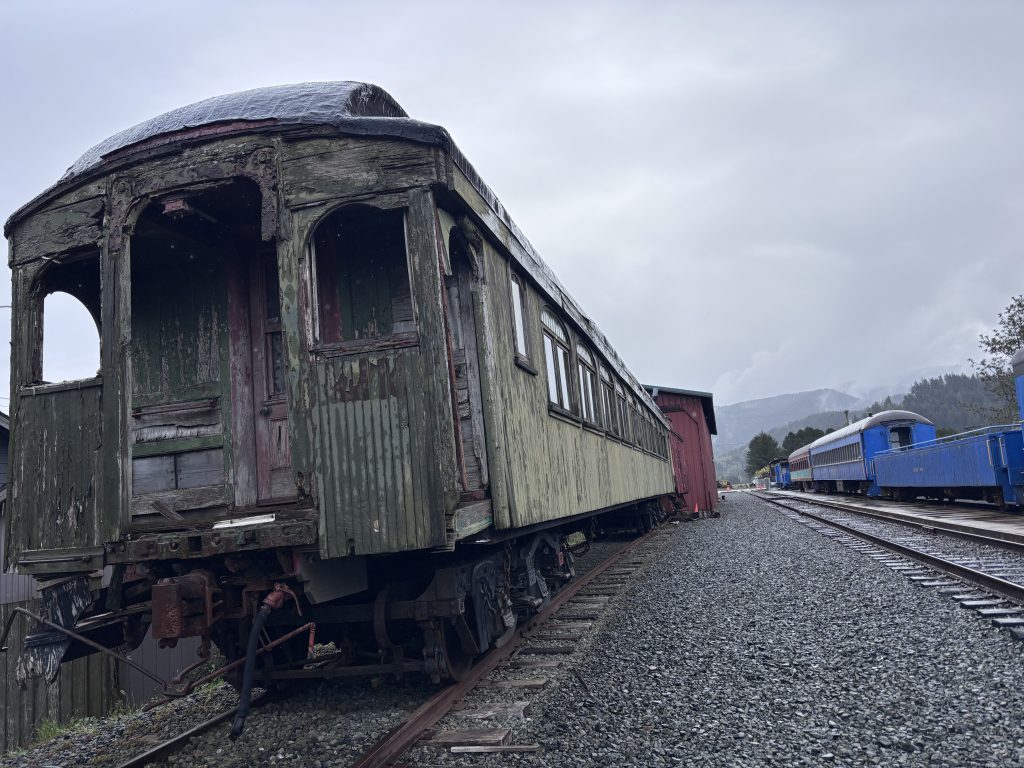 Aged green wooden passenger train car with rotting wood and missing panels on a gravel rail line, with blue and silver train cars visible in the background.