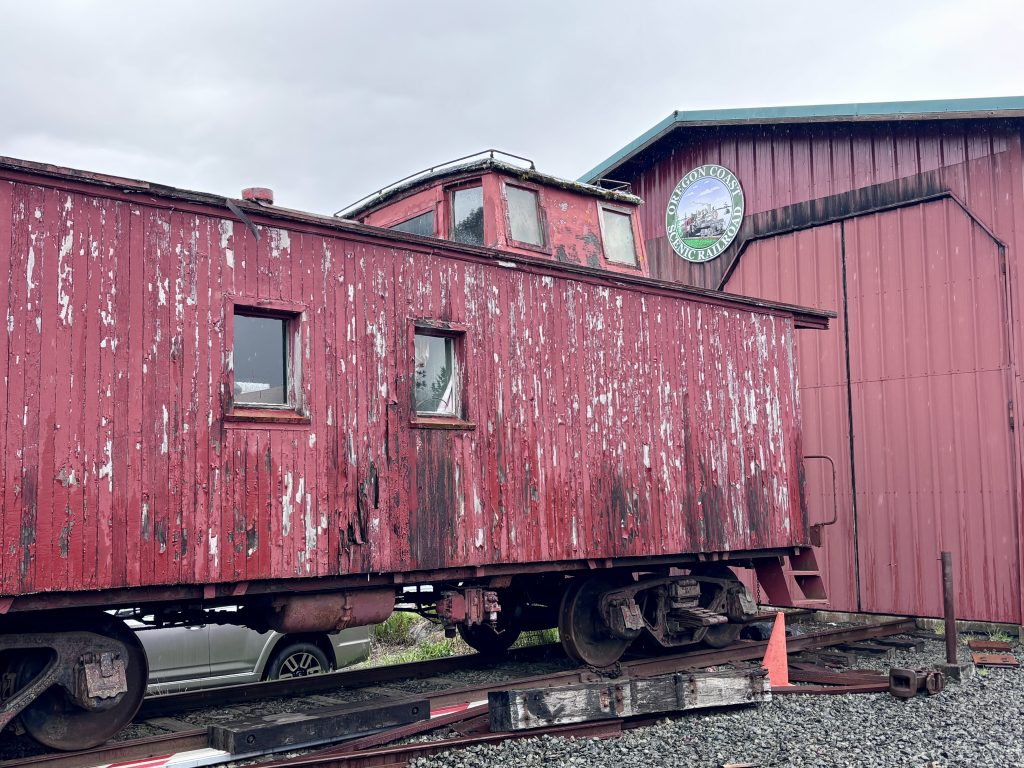 Weathered red caboose with peeling paint parked on old tracks beside the Oregon Coast Scenic Railroad building on an overcast day.