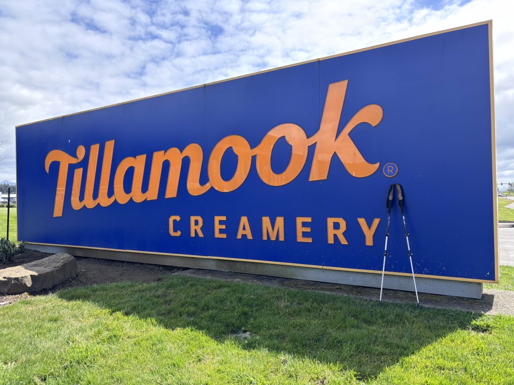 Large blue Tillamook Creamery sign with orange lettering and hiking poles leaning against it on a sunny day.