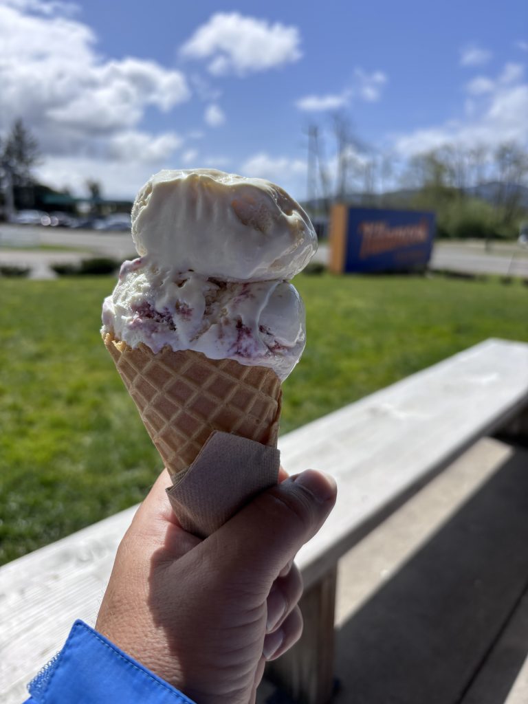 Hand holding a waffle cone with two scoops of Tillamook ice cream outdoors near the creamery under blue skies with scattered clouds.