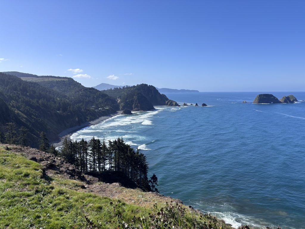Panoramic coastal view of cliffs, forested hills, and sea stacks along the Oregon Coast under bright blue skies.