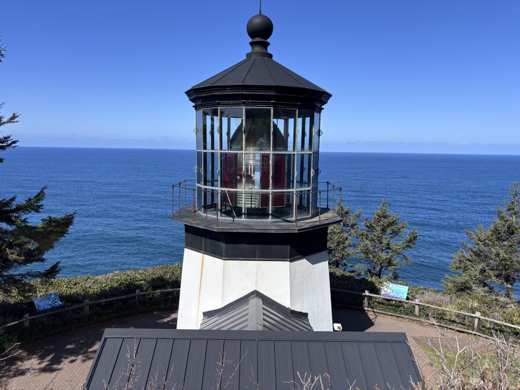 Historic lighthouse overlooking the Pacific Ocean, with a glass lantern room and red Fresnel lens against a clear blue sky at Cape Meares.