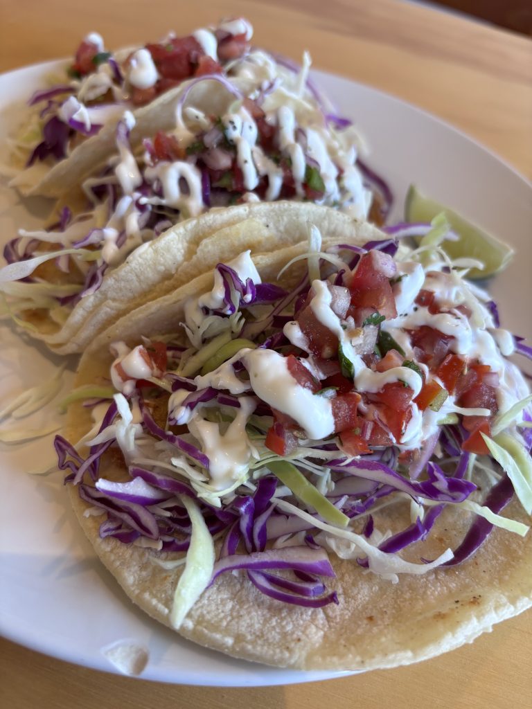Plate of fish tacos topped with shredded cabbage, diced tomatoes, and creamy white sauce served on soft corn tortillas.