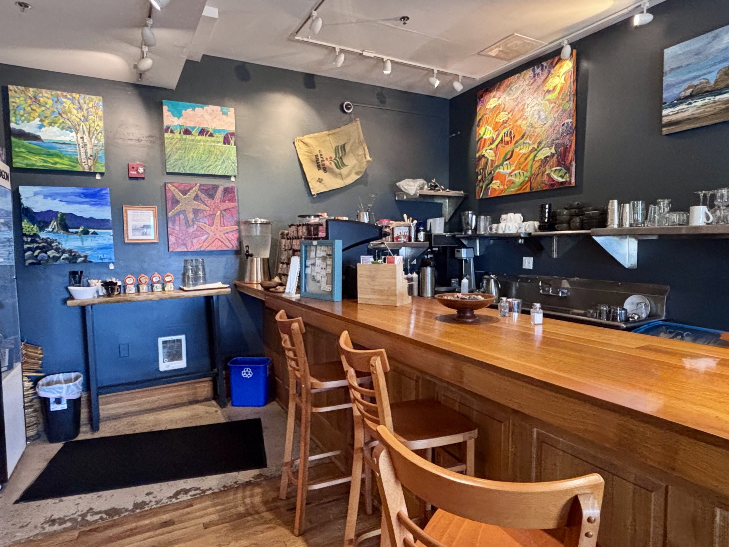 Cozy coastal café interior with a wooden bar, stools, espresso machine, and colorful artwork decorating dark gray walls.