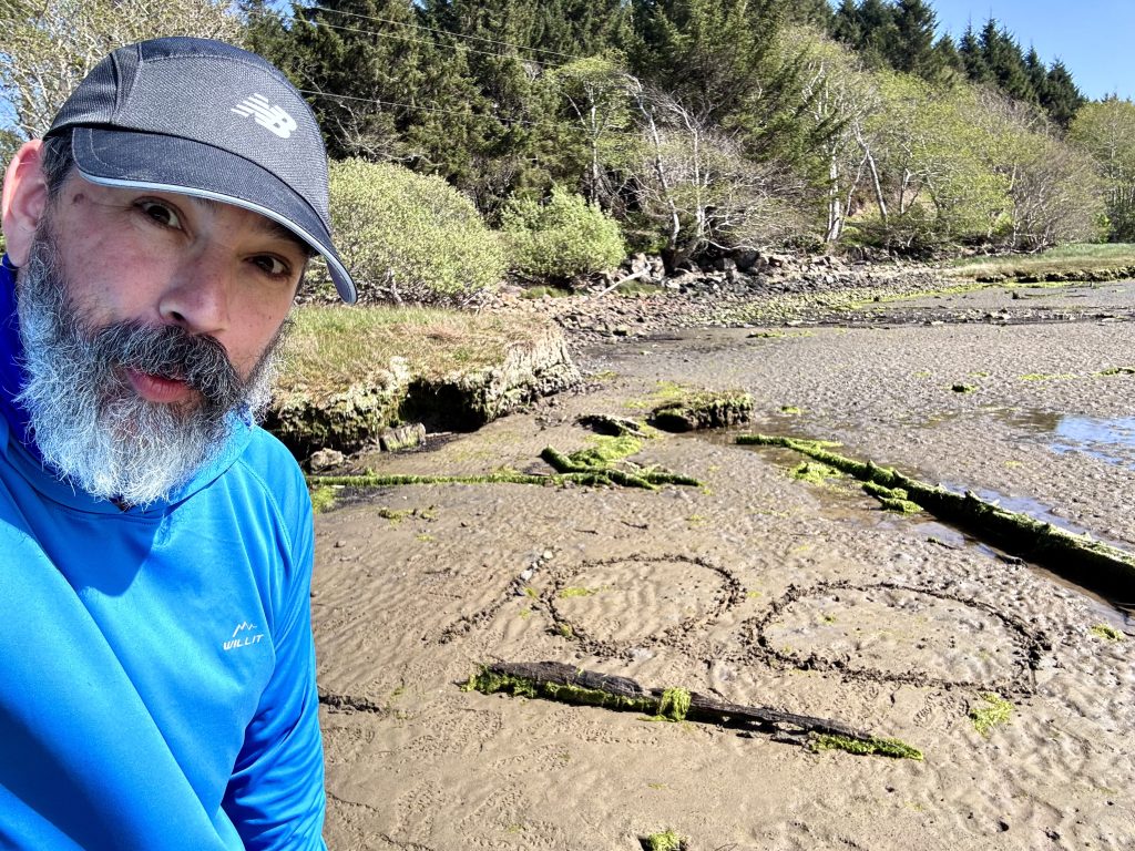 Man wearing a blue hiking shirt and black cap standing on a muddy shoreline with “100” drawn in the sand, surrounded by mossy logs and coastal trees.