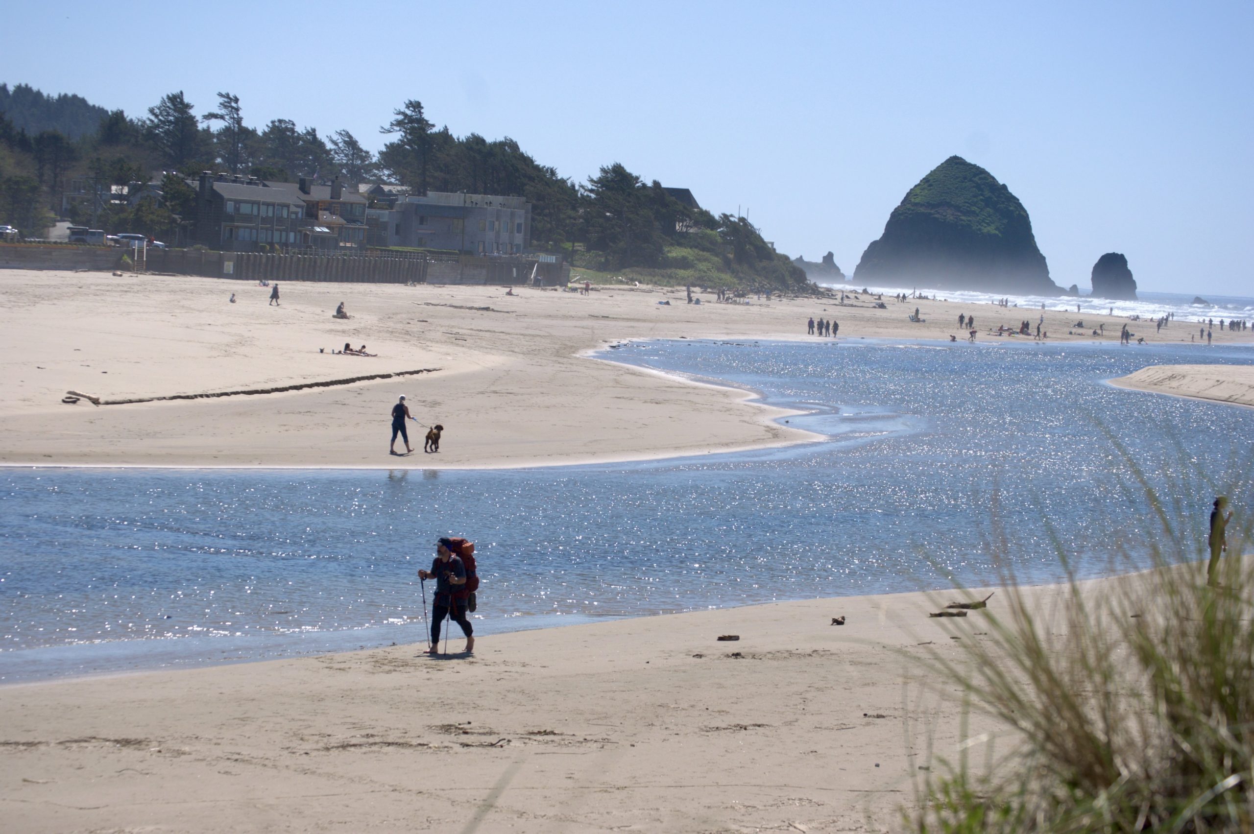 Backpacker crossing a sandy beach near Cannon Beach, Oregon, with a large red pack and trekking poles, while beachgoers and Haystack Rock are visible in the background across the reflective waters of Ecola Creek.