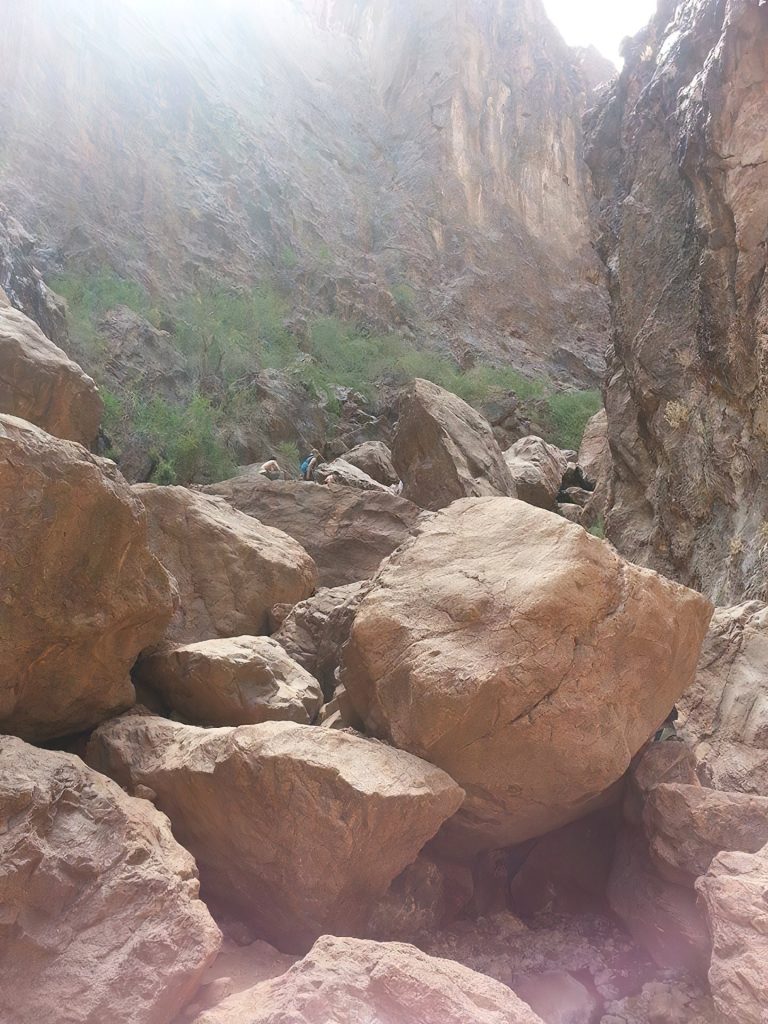 Large boulders in a rocky canyon with sunlight streaming in from above. Two people are climbing among the rocks.