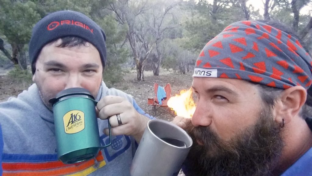 Two bearded men wearing knit caps drink from camping mugs in front of a small fire in a wooded outdoor setting.