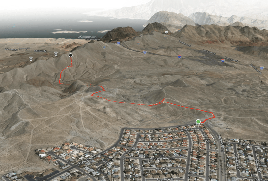 A 3D satellite map showing a red hiking route through desert hills near a residential area. The trail starts at a neighborhood street and winds through the rocky terrain toward a viewpoint overlooking Lake Mead.
