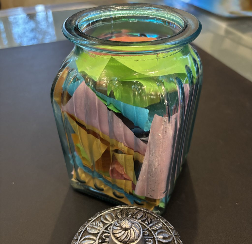Glass jar filled with colorful folded paper notes, with a decorative metal lid beside it.