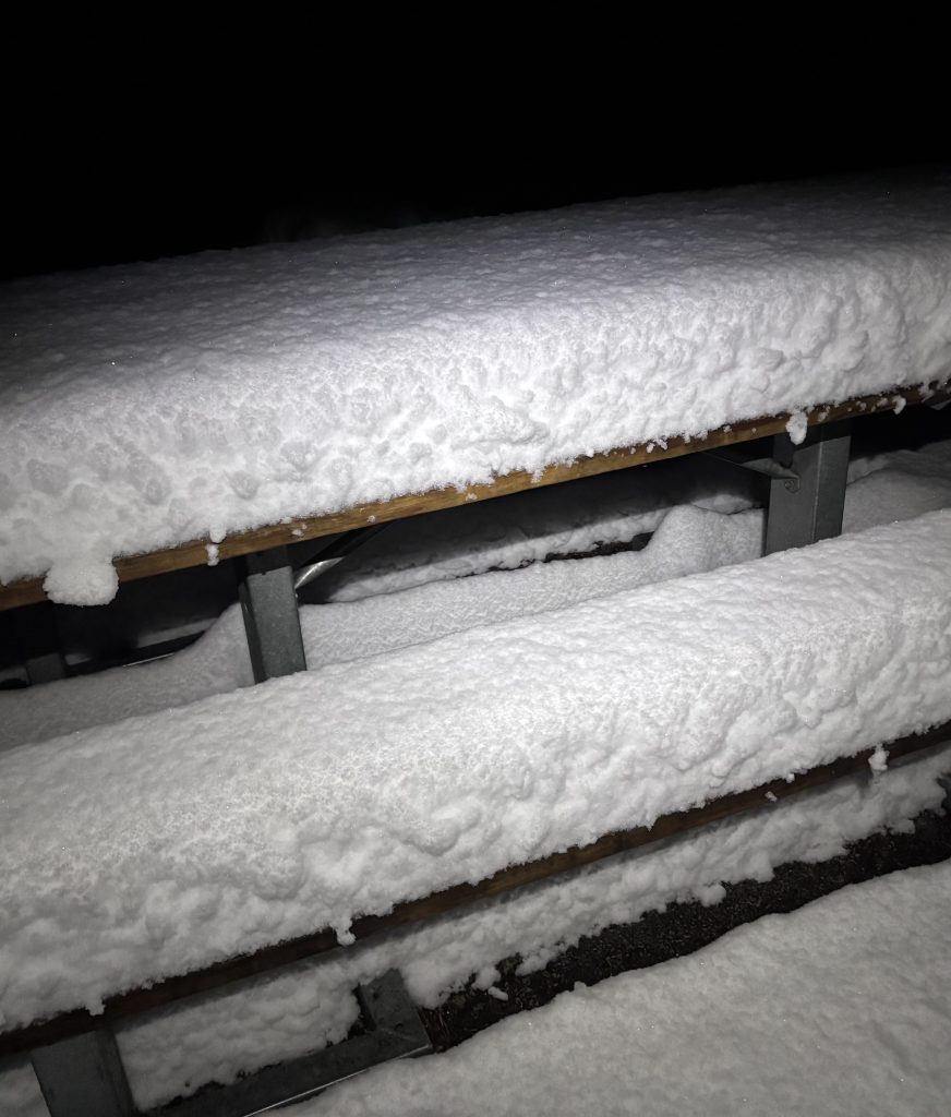 Wooden picnic table and benches covered in a thick layer of fresh snow at night.
