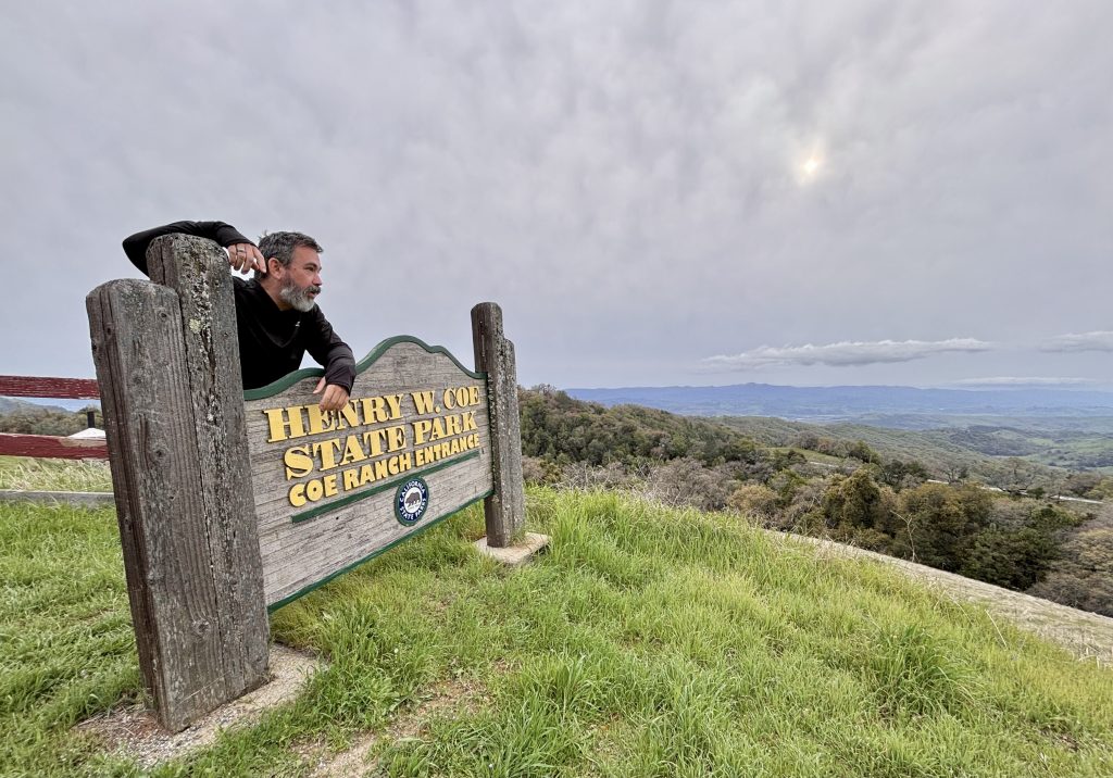 A middle-aged man stands behind the Entrance sign to Henry Coe State Park, with the Diablo range in the background, under a cloudy sky. 