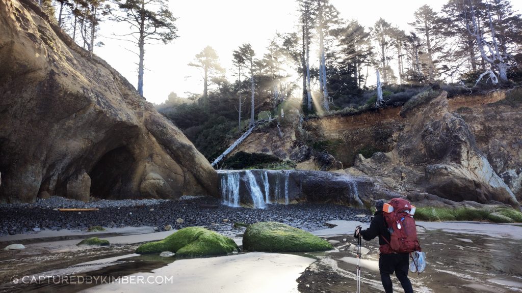 A scenic view of a beach featuring a small waterfall cascading down from a rocky cliff. A person wearing a backpack stands on the beach, looking towards the waterfall. The area is framed by dark cliffs on either side, with patches of green moss and foliage visible. A sandy beach stretches out toward the water.