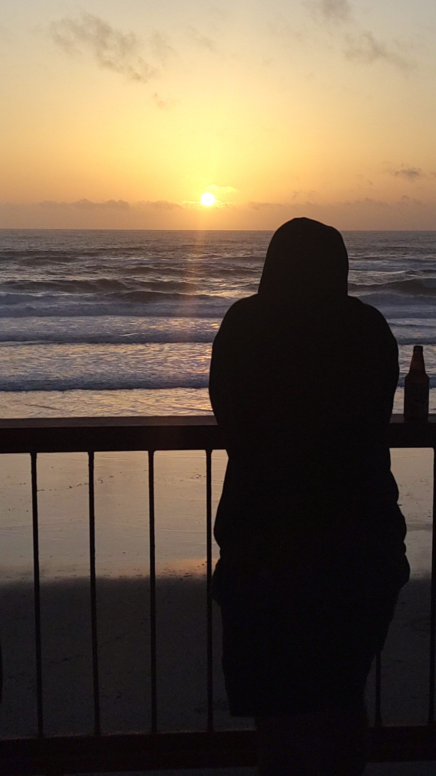 Silhouette of a person in a hoodie leaning against a beachside railing, watching the sun set over the Pacific Ocean. A bottle rests on the ledge beside them, waves rolling in below a golden sky.