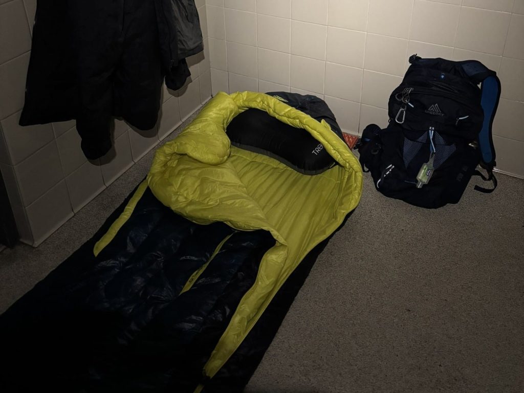 A yellow sleeping bag laid out on a hard floor with a black travel pillow inside; a dark backpack and hanging coats are nearby against a light-colored wall.