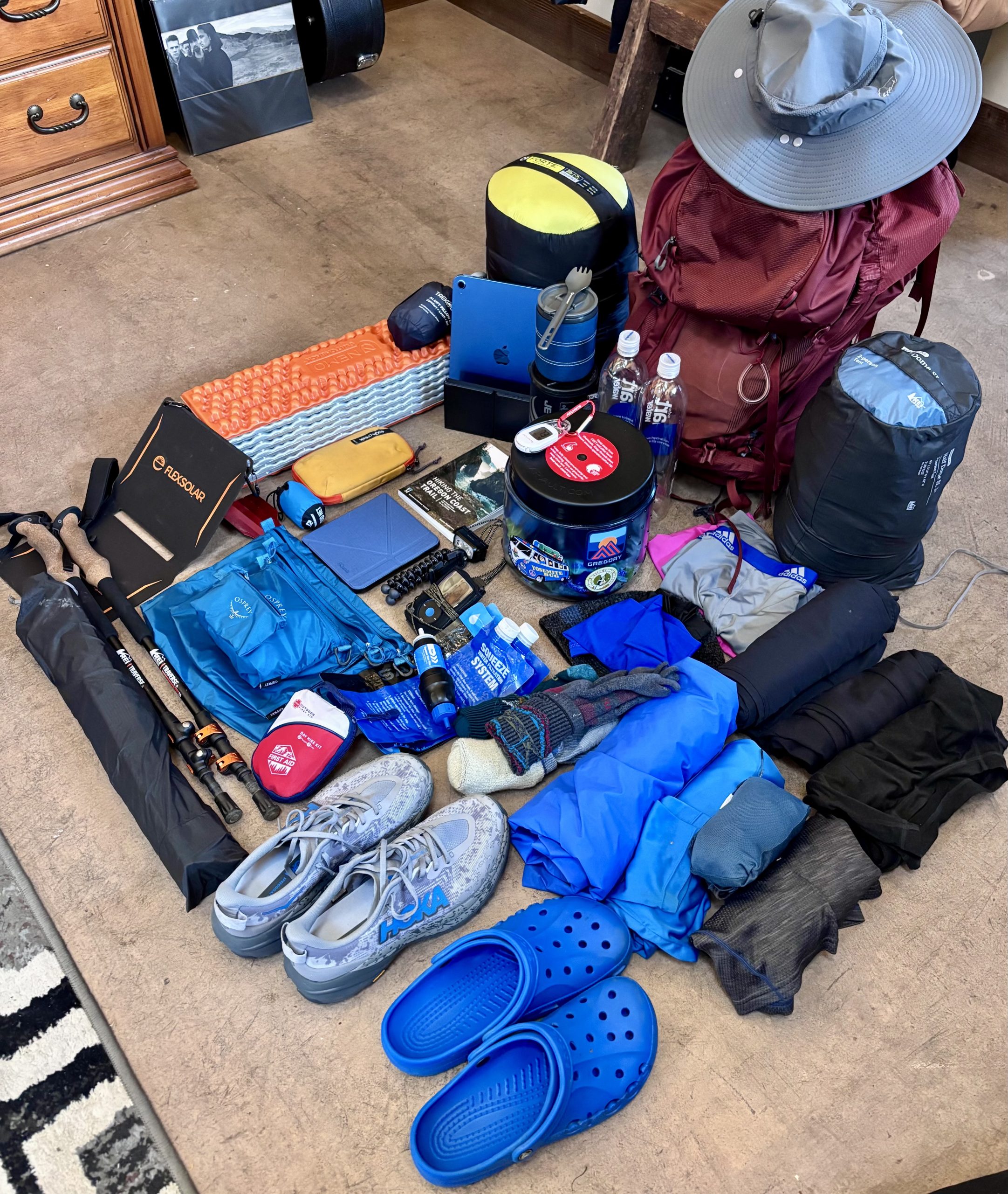 Overhead view of complete backpacking gear layout on carpeted floor, including a red Osprey hiking backpack with a wide-brim hat on top, gray HOKA trail running shoes, blue Crocs, bear canister, trekking poles, first aid kit, Therm-a-Rest Z Lite sleeping pad, electronics like an iPad and Kindle, two Smartwater bottles, sleeping bag, rain jacket, socks, headlamp, stove, utensils, compact umbrella, and clothing essentials — all meticulously arranged for a backpacking trip.