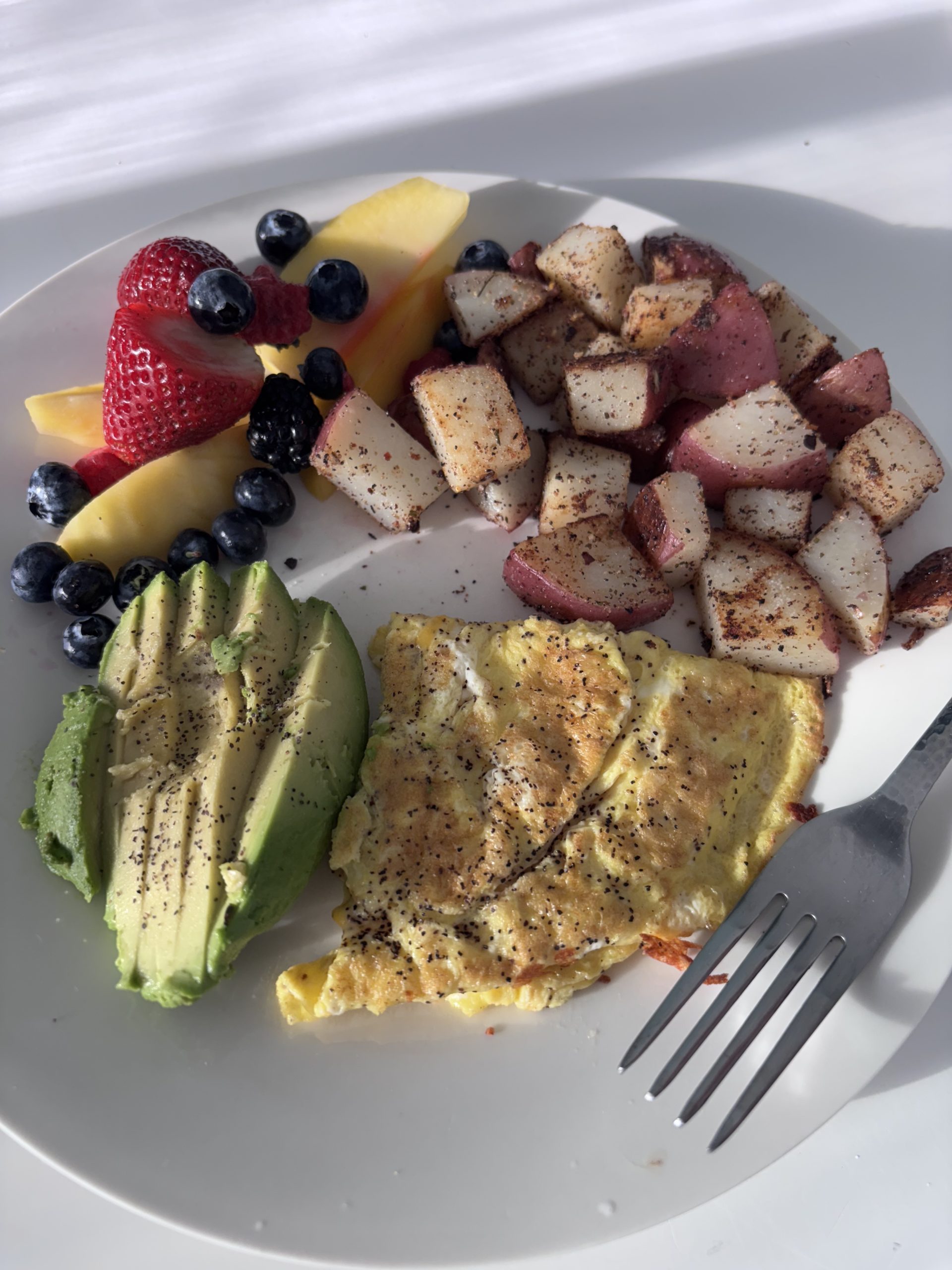 Healthy homemade breakfast with seasoned potatoes, avocado slices, scrambled eggs, and fresh fruit including strawberries, blueberries, and pineapple on a white plate.