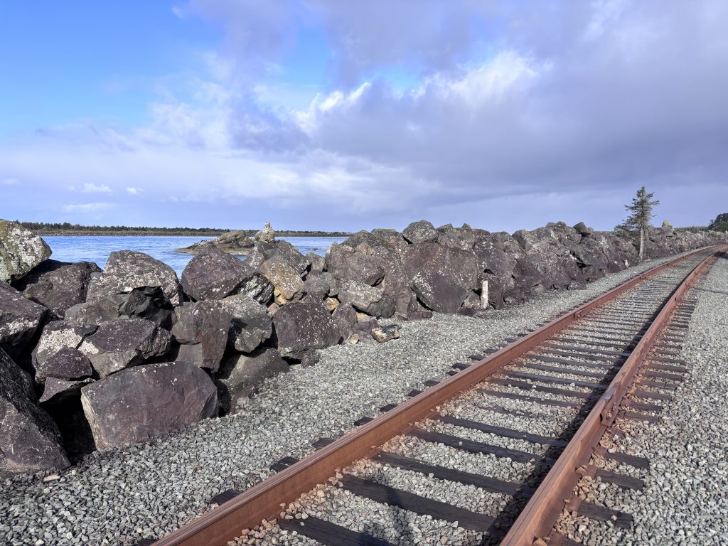 Rusty railroad tracks curving beside large black boulders along the water’s edge near the mouth of a bay on the Oregon Coast.