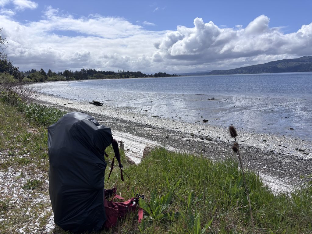 Backpack with rain cover resting beside a rocky shoreline along Tillamook Bay under dramatic clouds and blue sky.