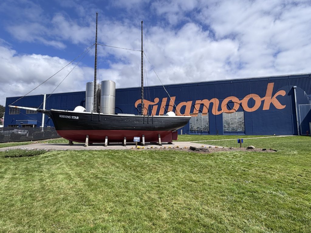 Historic wooden ship named Morning Star displayed outside the Tillamook Creamery building with large orange Tillamook lettering on a blue wall under partly cloudy skies.