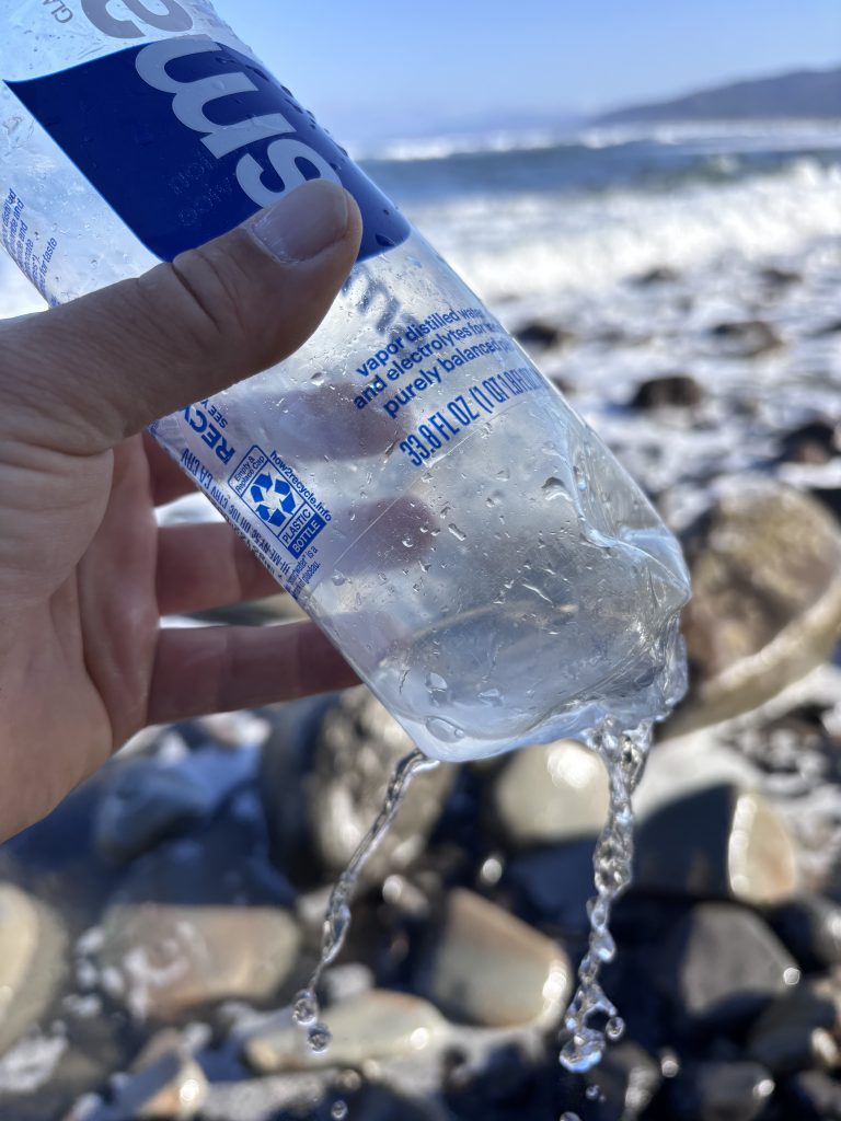 Hand holding a punctured Smartwater bottle with water pouring out, ocean waves and rocky shoreline blurred in the background.