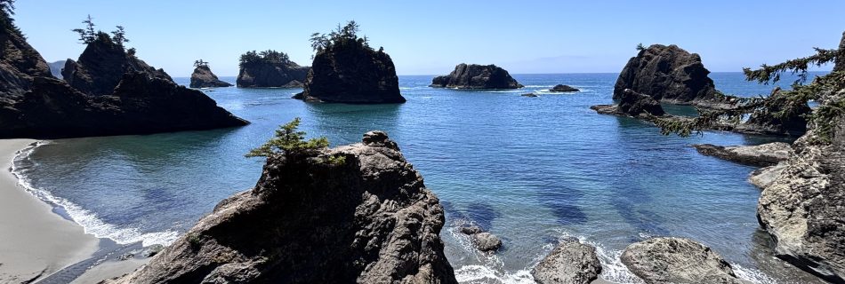 Wide-angle view of the rugged Oregon Coast at Secret Beach, showcasing sea stacks rising from turquoise water under a bright blue sky, bordered by cliffs and sandy coves.